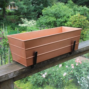 May include: A copper-colored metal planter box with black metal brackets, mounted on a wooden railing. The planter is designed for hanging on a railing.