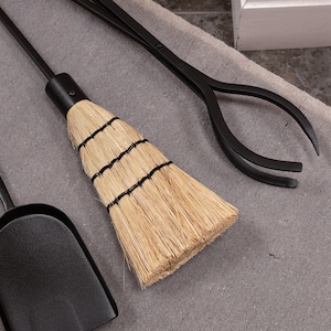 May include: A set of black fireplace tools, including a shovel, tongs, and a small broom. The broom has a natural straw head bound with black twine. The tools are arranged on a gray surface.