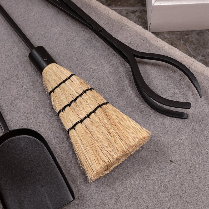 May include: A set of black fireplace tools, including a shovel, tongs, and a small broom. The broom has a natural straw head bound with black twine. The tools are arranged on a gray surface.