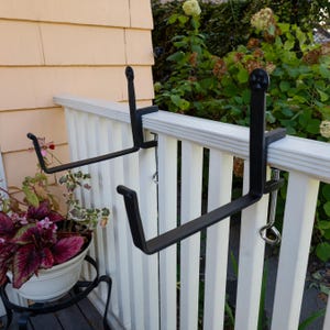 May include: Two black metal plant brackets mounted on a white wooden railing. The brackets are designed to hold flower pots.
