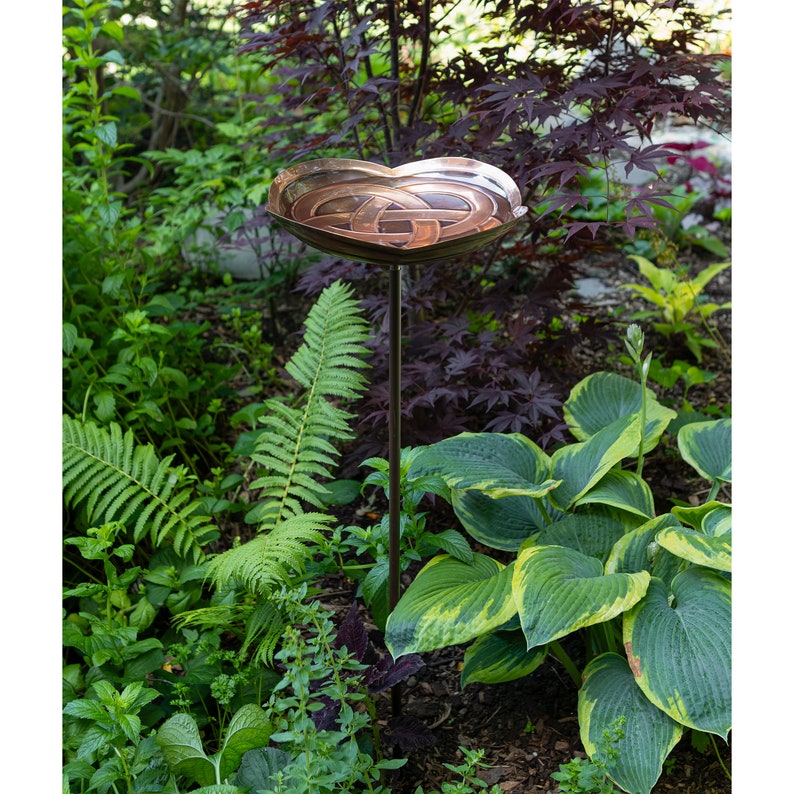 K&ouml;nnte beinhalten: Ein Vogelbad aus Kupfer mit einem keltischen Knotenmuster steht auf einem hohen Metallst&auml;nder in einem Garten. Das Bad ist herzf&ouml;rmig und von &uuml;ppigen gr&uuml;nen Farnen und Hosta-Pflanzen umgeben.