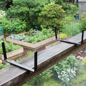 May include: Two black metal plant brackets mounted on a wooden fence rail. The brackets are designed to hold a plant pot or planter.
