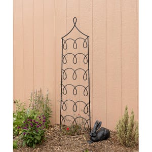 May include: Black metal garden trellis with a decorative design. The trellis is standing in a garden setting with green plants and a small black rabbit statue.