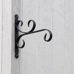 May include: Black wrought iron hanging basket bracket with decorative scrollwork. The bracket is mounted on a white wooden surface. The bracket has a vertical mounting plate and a curved arm for holding a hanging basket.
