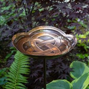 K&ouml;nnte beinhalten: Ein Vogelbad aus Kupfer mit einem keltischen Knotenmuster. Das Bad ist auf einem schwarzen Metallpfosten montiert und von gr&uuml;nem Laub umgeben.