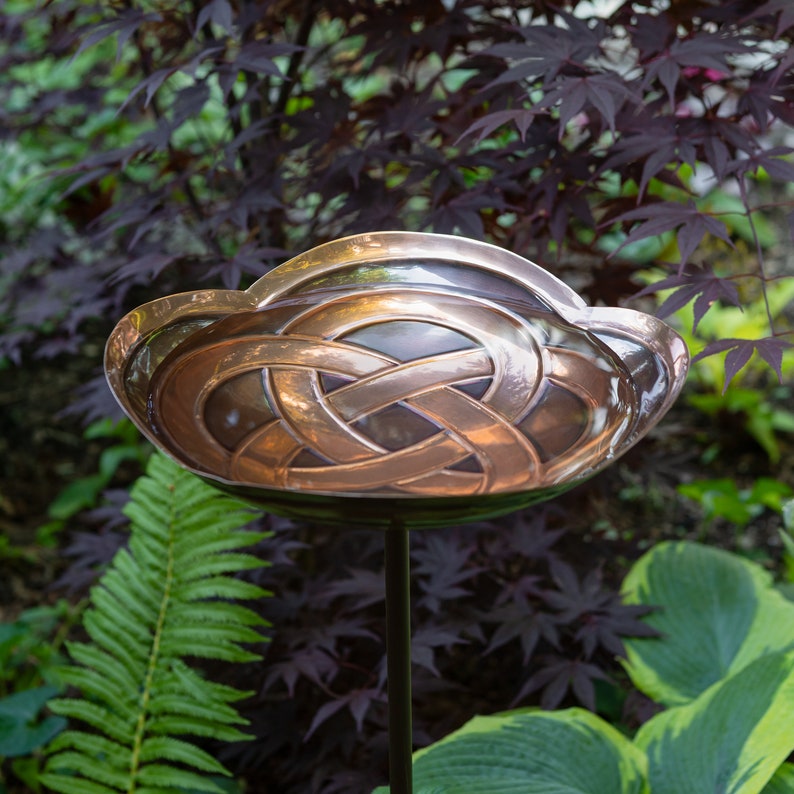 K&ouml;nnte beinhalten: Ein Vogelbad aus Kupfer mit einem keltischen Knotenmuster. Das Bad ist auf einem schwarzen Metallpfosten montiert und von gr&uuml;nem Laub umgeben.
