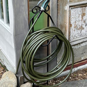 May include: A green garden hose coiled on a black metal holder, with a spray nozzle attached. The hose is next to a weathered wooden door and a window. The nozzle is white and teal. The scene is outdoors.
