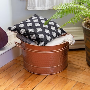 May include: A copper-colored metal storage bin with a hammered texture, holding a black and white patterned pillow and folded blankets. The bin has two handles and sits on a wooden floor. A potted fern is in the background.