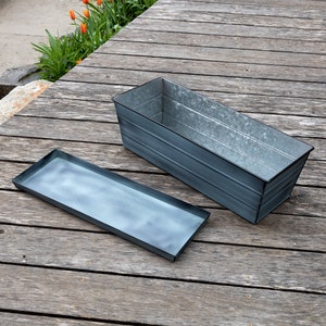 May include: A galvanized metal planter with a matching tray. The planter is rectangular and has a rustic, weathered look. The tray is also rectangular and has a slightly raised edge.