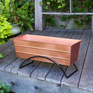 May include: A copper planter with a black metal stand. The planter is rectangular and has a slightly rounded edge. The stand is made of two curved pieces of metal that meet in the middle.