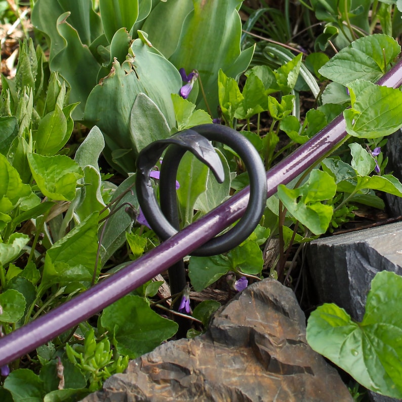 Puede incluir: Una estaca de jard&iacute;n de metal negro con un dise&ntilde;o decorativo de espiral. La estaca se inserta en el suelo y tiene un acabado pintado de color morado.