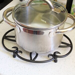 Op de afbeelding: Een zilveren pot met een glazen deksel staat op een zwarte metalen onderzetter. De pot heeft twee handvatten en staat op een wit aanrecht.