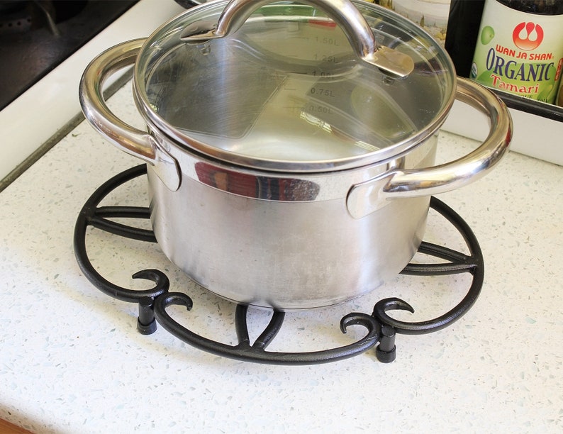 Op de afbeelding: Een zilveren pot met een glazen deksel staat op een zwarte metalen onderzetter. De pot heeft twee handvatten en staat op een wit aanrecht.