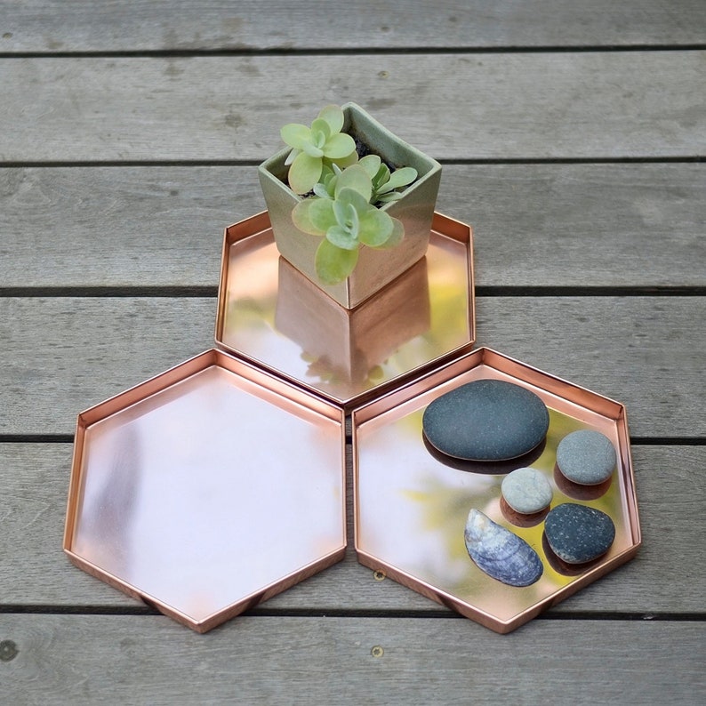 May include: Three copper-coloured hexagonal trays on a wooden surface. One tray holds a small square ceramic planter with a succulent. The other trays contain smooth stones and a seashell. The trays have a reflective surface.