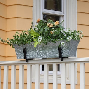 May include: A silver metal planter with a decorative floral pattern is mounted on a white railing. The planter is filled with a variety of green plants and flowers, including white and orange blooms.