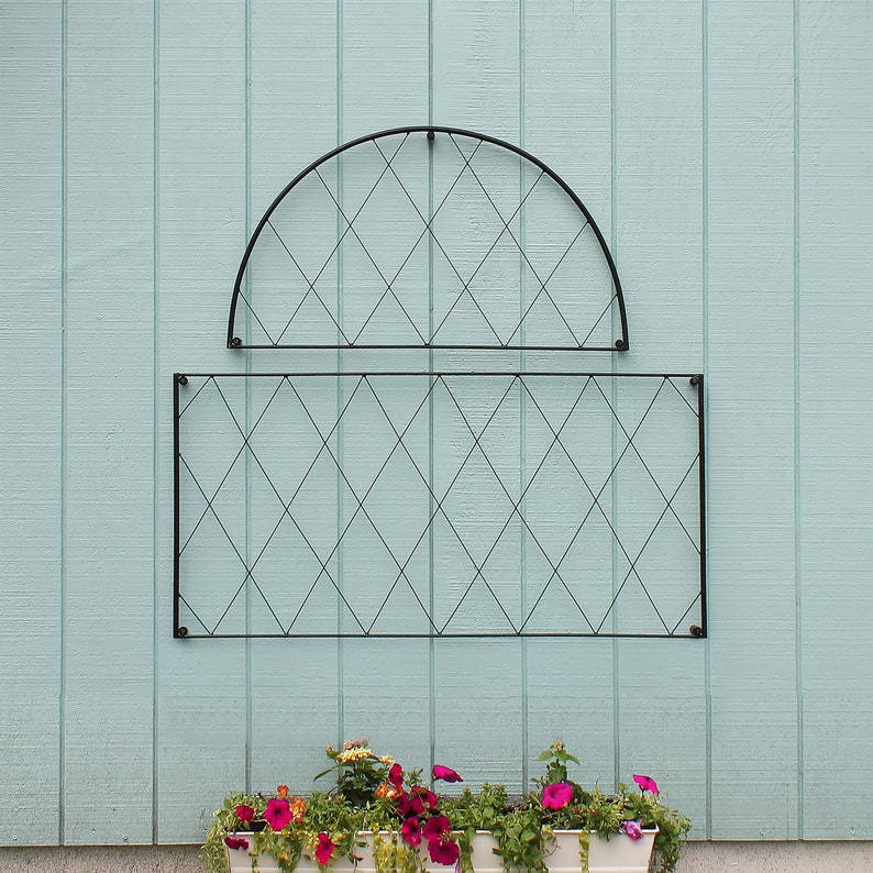 Op de afbeelding: Zwart metalen trellis met een ruitpatroon, bevestigd aan een lichtblauwe muur. De trellis is in twee delen, een gebogen en een rechthoekig deel. Een plantenbak met roze bloemen staat voor de trellis.