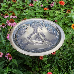 May include: A metal bird bath with a design of three hares in a circle. The bath is on a black metal stand and is surrounded by green plants and flowers.