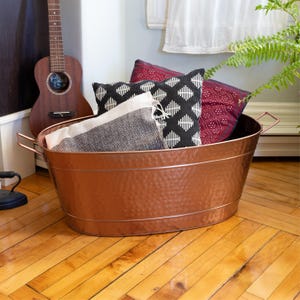 May include: A hammered copper oval tub with two handles, filled with pillows and a blanket. The pillows have geometric patterns in black, white, and burgundy. A guitar leans against the wall in the background.