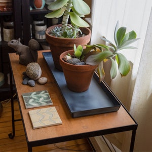 May include: A black metal table with a wooden top. Two potted plants are on the table, one with green leaves and the other with gray-green leaves. A small squirrel figurine is on the table.