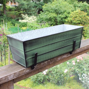 May include: A green metal planter box with black metal brackets. The planter is mounted on a wooden railing.