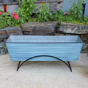 May include: A light blue metal planter with a black metal stand. The planter is rectangular and has a slightly curved top. The stand has two curved legs.