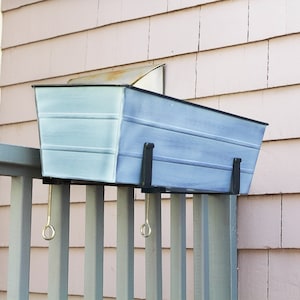 May include: A light blue metal planter with black metal brackets attached to a gray wooden railing. The planter is hanging from the railing.