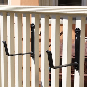May include: Two black metal plant hangers mounted on a white wooden fence. The hangers have a decorative scroll design at the top and a curved arm for supporting a plant pot.