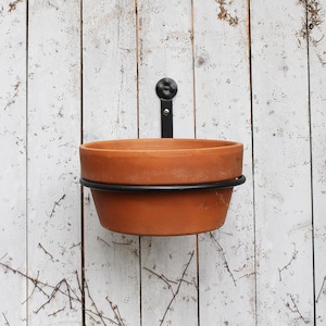 May include: A terracotta planter with a black metal ring and hook for hanging on a white wooden wall.