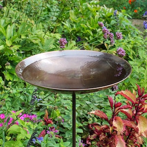 May include: A hammered copper bird bath with a wide, shallow bowl and a dark metal stand. The bird bath is filled with water and surrounded by lush green foliage, purple flowers, and red and green plants.