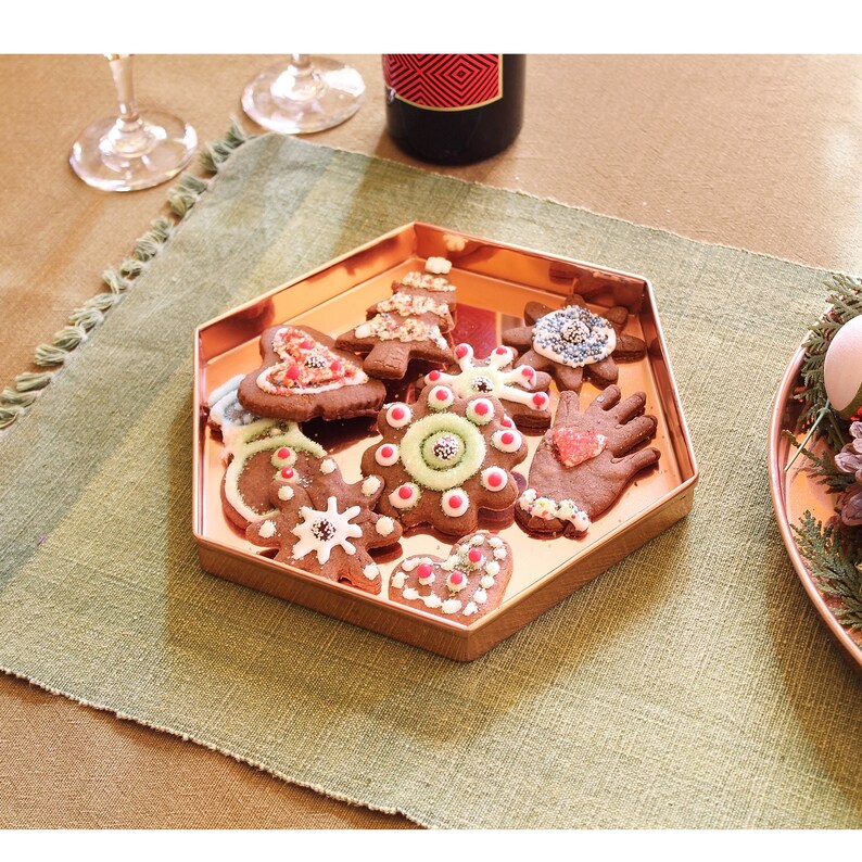 May include: A copper-coloured, hexagonal tray filled with decorated gingerbread biscuits. The biscuits are in various shapes, including trees, snowflakes, and hearts, and are adorned with white icing and colourful sprinkles. The tray sits on a green placemat.