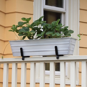 May include: White metal planter box with green plants growing inside, mounted on a white wooden railing with black metal brackets.