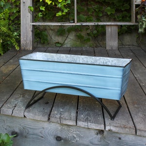 May include: A light blue metal planter with a black metal stand. The planter has a rectangular shape and is sitting on a wooden surface.