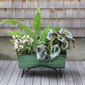 May include: A green metal planter with a black metal stand, filled with ferns and white flowers. The planter is sitting on a wooden deck.