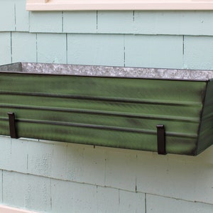 May include: A green metal planter box with a distressed finish, mounted on a light blue wall. The planter has two black metal brackets.