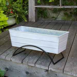 May include: White metal planter with a black metal stand. The planter is rectangular and has a slightly distressed finish. The stand is a simple arch shape.