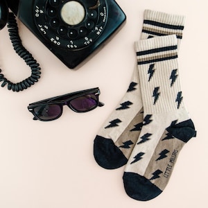 May include: A pair of beige socks with black lightning bolt patterns and navy blue accents. The socks have a striped design at the top and the words "LITTLE MILLERS" at the bottom. Black sunglasses and a vintage telephone are also in the image.