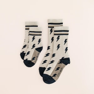 May include: Four pairs of crew socks with a light beige base color. Each sock features a pattern of black lightning bolts and a navy blue heel and toe. The top of the socks have a striped design in black, beige, and brown.