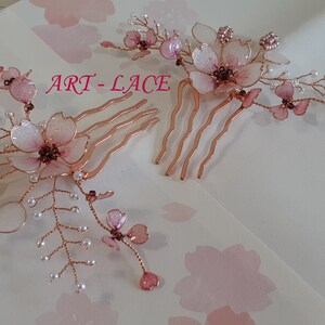 May include: Two hair combs featuring rose gold wire branches adorned with translucent pink cherry blossom petals and small white beads. The text ART-LACE is printed in pink between the two combs, which are arranged on a light surface.