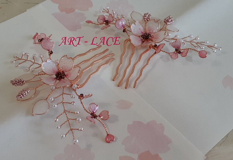 May include: Two hair combs featuring rose gold wire branches adorned with translucent pink cherry blossom petals and small white beads. The text ART-LACE is printed in pink between the two combs, which are arranged on a light surface.