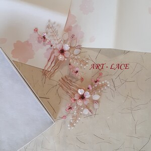 May include: Two hair combs featuring rose gold wire branches adorned with translucent pink flower petals, small white beads, and dark pink accents. The text ART-LACE is visible in red. The design mimics delicate floral sprays with metallic stems.