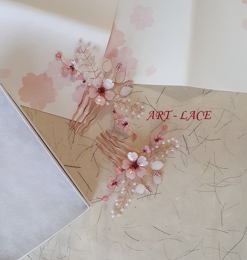 May include: Two hair combs featuring rose gold wire branches adorned with translucent pink flower petals, small white beads, and dark pink accents. The text ART-LACE is visible in red. The design mimics delicate floral sprays with metallic stems.