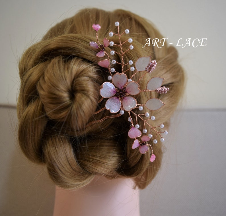 May include: A hair comb featuring delicate copper wire branches adorned with translucent pink and white flower petals, small white beads, and dark centers. The design includes the text ART-LACE. The floral arrangement is secured in a braided hairstyle.