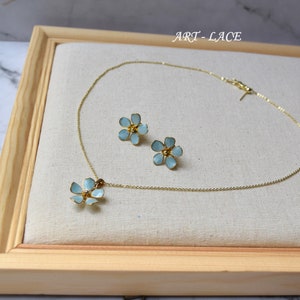May include: A gold chain necklace with a single light blue flower pendant and a pair of matching light blue flower stud earrings. The jewelry set features gold-toned metal edges and the text ART-LACE is visible in the upper right corner.