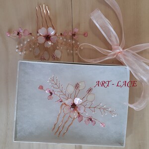May include: Two hair combs featuring rose gold wire, translucent pink flower petals, and small white pearl-like beads. One comb rests inside a white box labeled ART-LACE, while the other sits above it next to a sheer light pink ribbon.