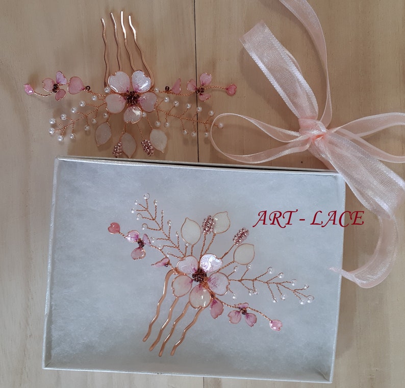 May include: Two hair combs featuring rose gold wire, translucent pink flower petals, and small white pearl-like beads. One comb rests inside a white box labeled ART-LACE, while the other sits above it next to a sheer light pink ribbon.