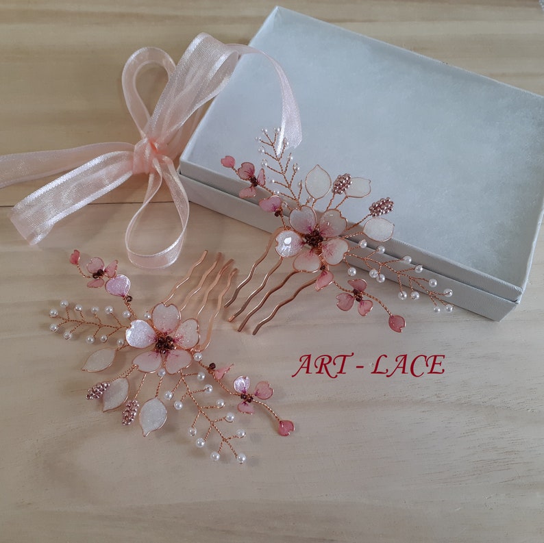 May include: Two rose gold hair combs featuring delicate wire-wrapped floral designs with translucent pink petals, small white beads, and metallic accents. A light pink ribbon is attached, and the text ART-LACE is printed in red on the surface.