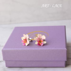 Può includere: Orecchini a forma di fiori di orchidea con petali smaltati bianchi, rosa e arancioni. Gli orecchini sono montati su perni d'argento e poggiano su una scatola lilla. Il testo "ART - LACE" è visibile.