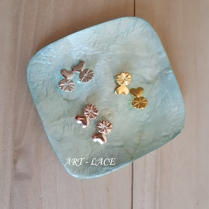 Op de afbeelding: Een lichtblauw vierkant bord met een getextureerd oppervlak. Drie sets gouden hartvormige metalen versieringen met een bloemmotief zijn op het bord gerangschikt. De tekst "ART - LACE" is op het bord gedrukt.