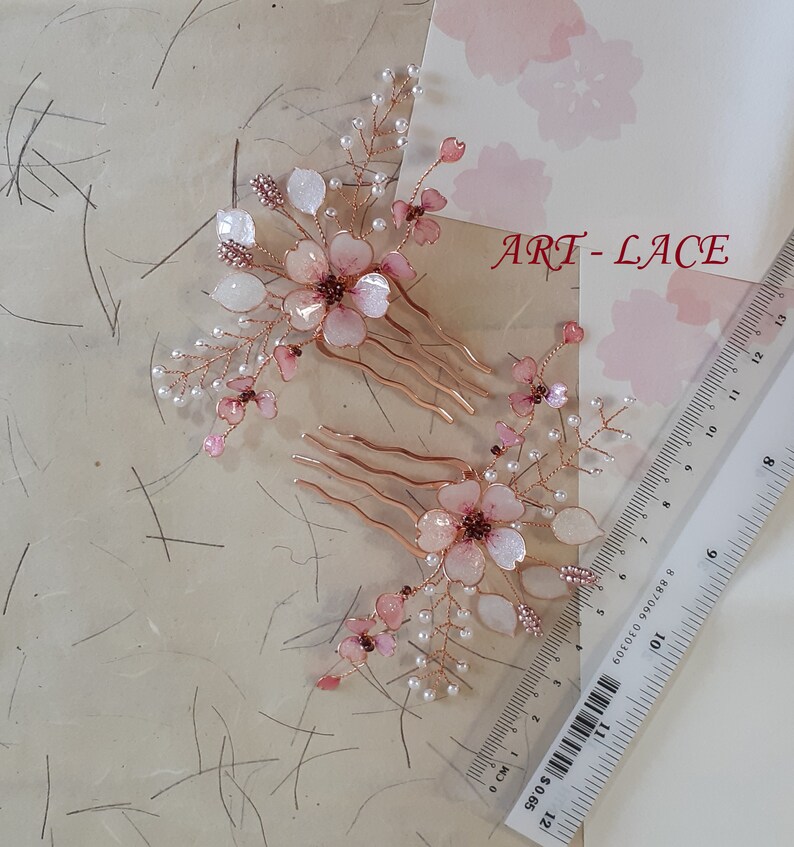 May include: Two hair combs featuring rose gold wire branches adorned with translucent pink and white flower petals and small white beads. A clear plastic ruler shows the combs measure approximately 4 inches in length. The text ART-LACE is visible.