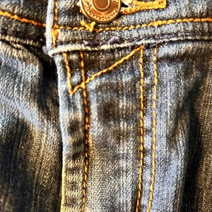 May include: Close-up of denim jeans with a button and orange stitching. The button is a copper color with the word "Seven" around the edge. The jeans have a classic five-pocket design.